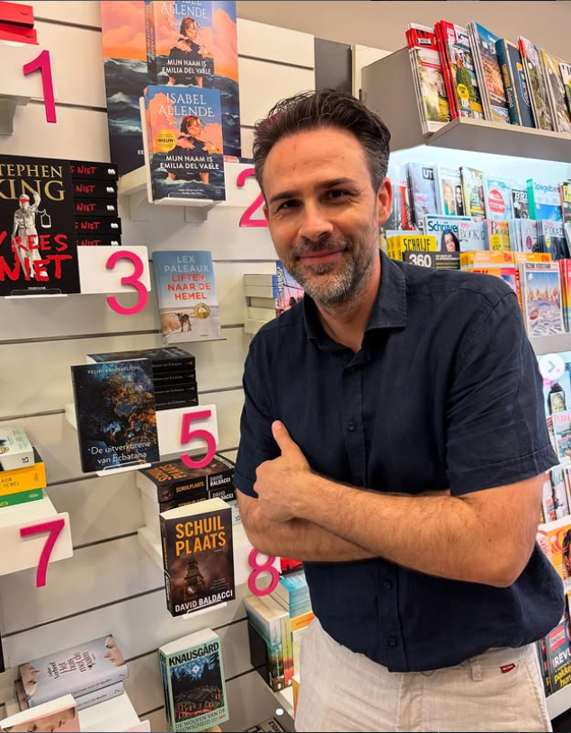 Veljko Radosavljevic en zijn boek "De uitverkorene van Ecbatana" Een middeljarige man staat trots in een boekwinkel, zijn eigen boek staat in de kast, op nr 5 van een reeks uitgelichte boeken.